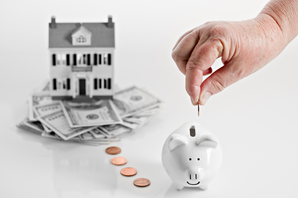 A conceptual image showing a reverse mortgage with a home sitting on a pile of money and a hand dropping coins slowly in a piggy bank. PLEASE CLICK HERE IF YOU NEED MORE MONEY