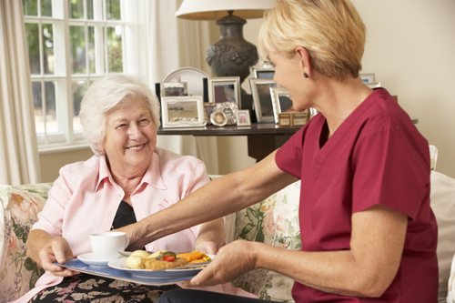 Helper Serving Happy Senior Woman With Meal In Care Home.
