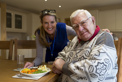 Community care worker serves up lunch to an elderly man sitting at the kitchen table.