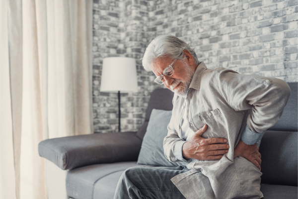 Older adult man sitting on sofa holding side due to back pain