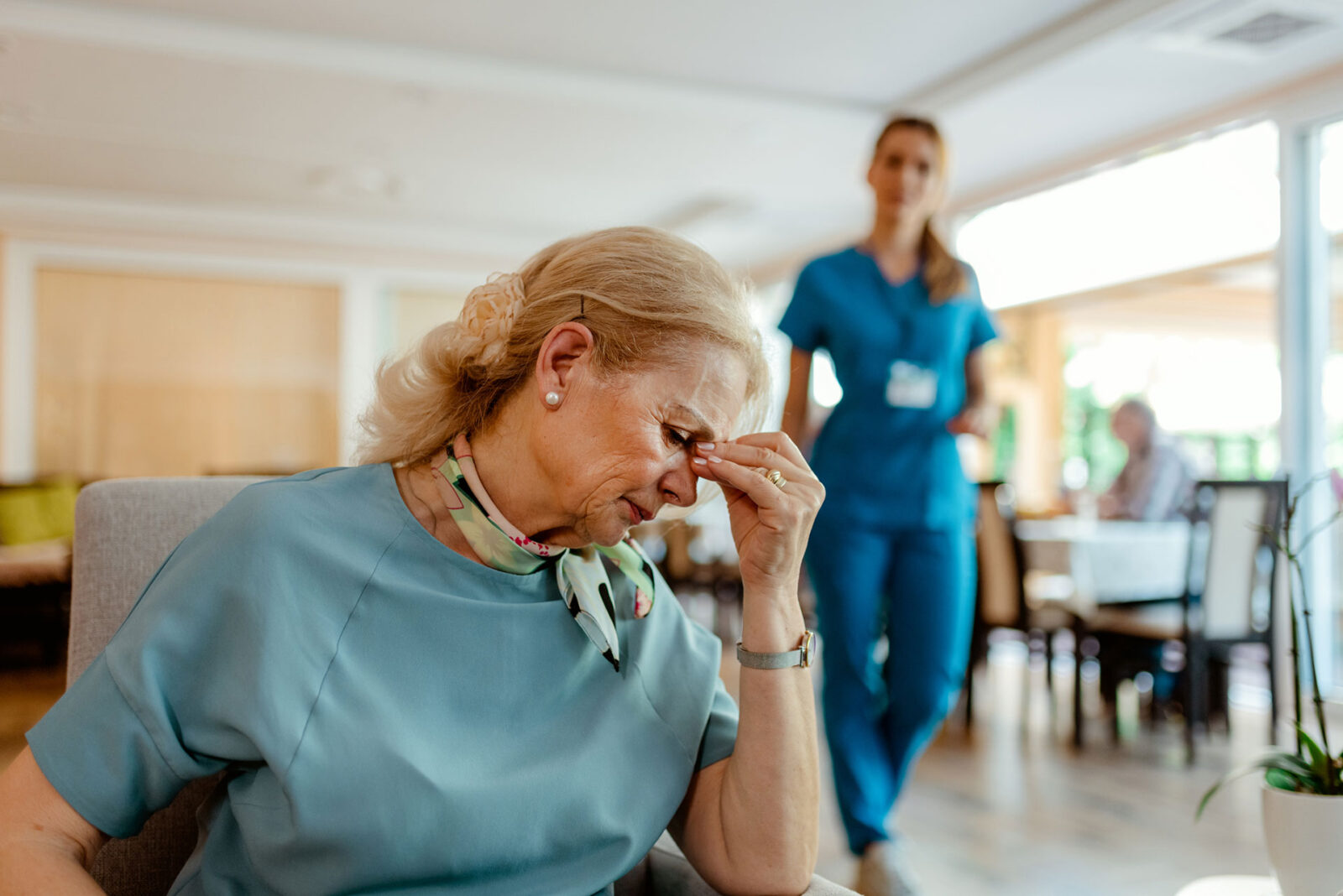 An elderly woman looks distressed, a caregiver approaches her.